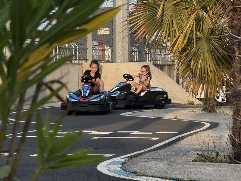 Deux enfants souriants conduisent des karts sur la piste, entourés de végétation et palmiers.