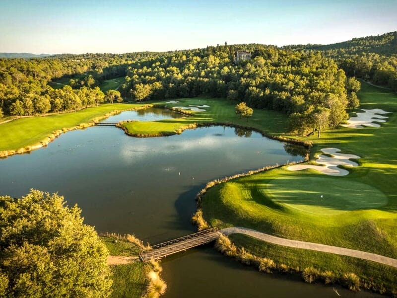 Vue aérienne d'un parcours de golf avec un lac, un pont en bois et des zones de sable entourées d'arbres verts.