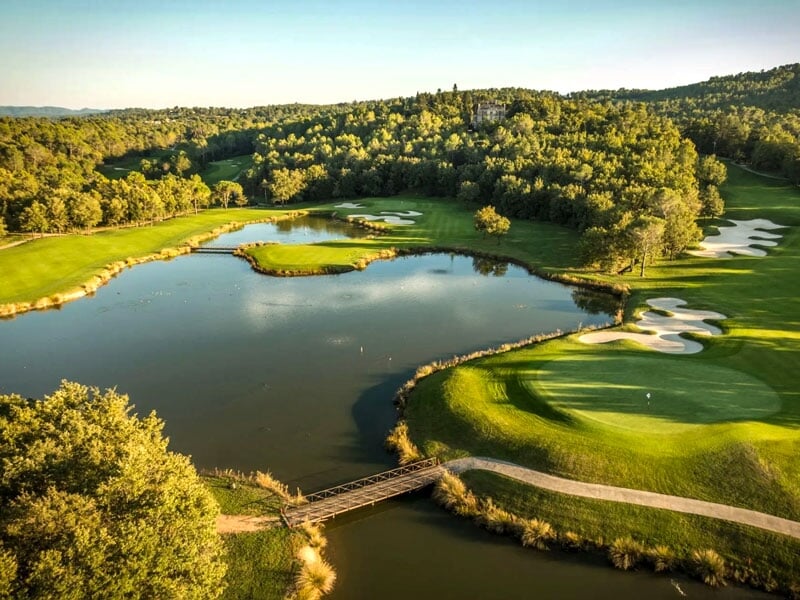 Vue aérienne d'un parcours de golf avec un lac, un pont en bois et des zones de sable entourées d'arbres verts.