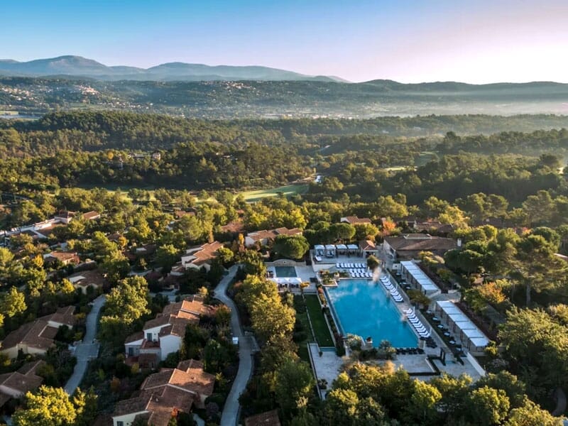 Vue panoramique d'un complexe hôtelier avec une grande piscine, des bâtiments et une forêt dense en arrière-plan.
