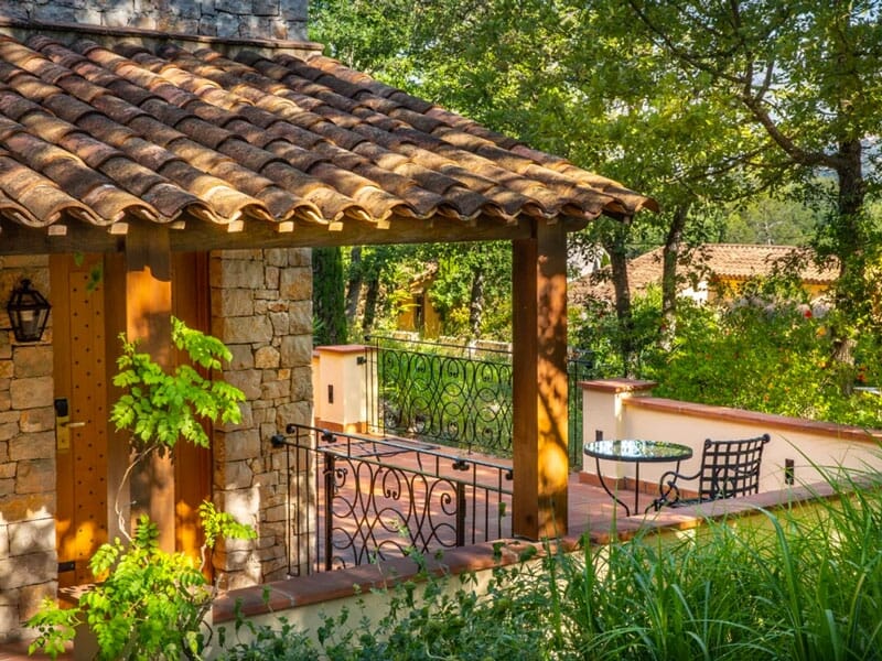 Terrasse d'une maison en pierre avec un toit en tuiles, une table ronde et des chaises entourées de verdure.