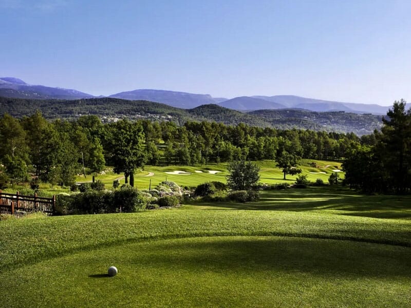Vue d'un terrain de golf avec un départ, des bunkers et une forêt dense sous un ciel clair et ensoleillé.