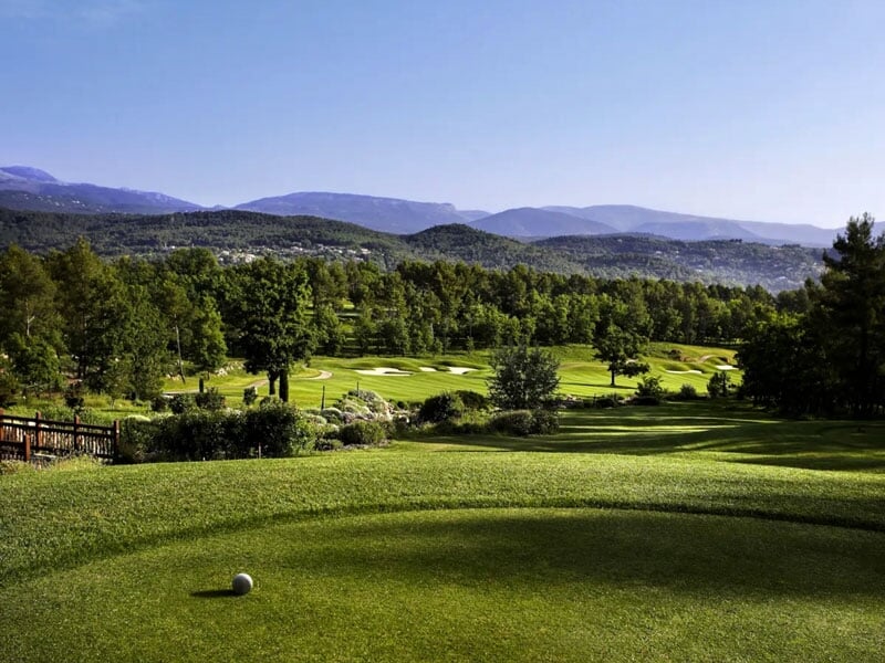 Vue d'un terrain de golf avec un départ, des bunkers et une forêt dense sous un ciel clair et ensoleillé.