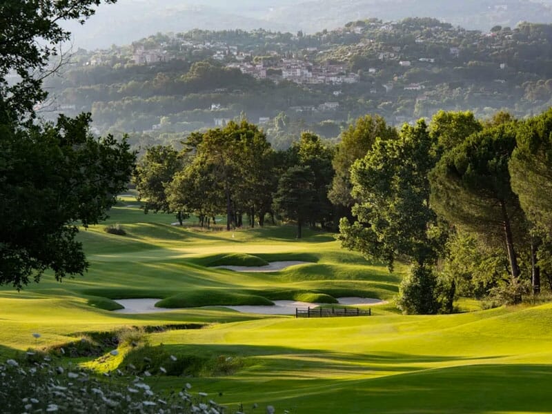 Paysage vallonné d'un parcours de golf avec des fairways verts, des bunkers et des arbres sous un ciel bleu.