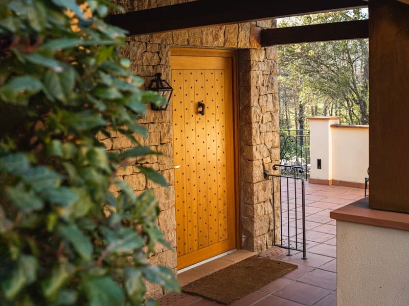 Entrée d'une maison en pierre avec une porte en bois ornée, un petit portail et des plantes autour.
