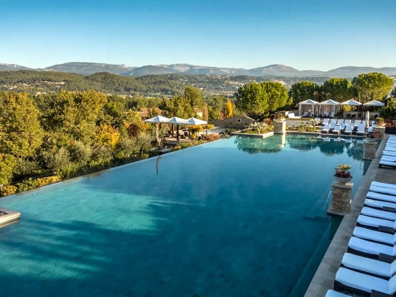 Piscine à débordement avec des chaises longues blanches, entourée d'arbres et offrant une vue sur les collines.