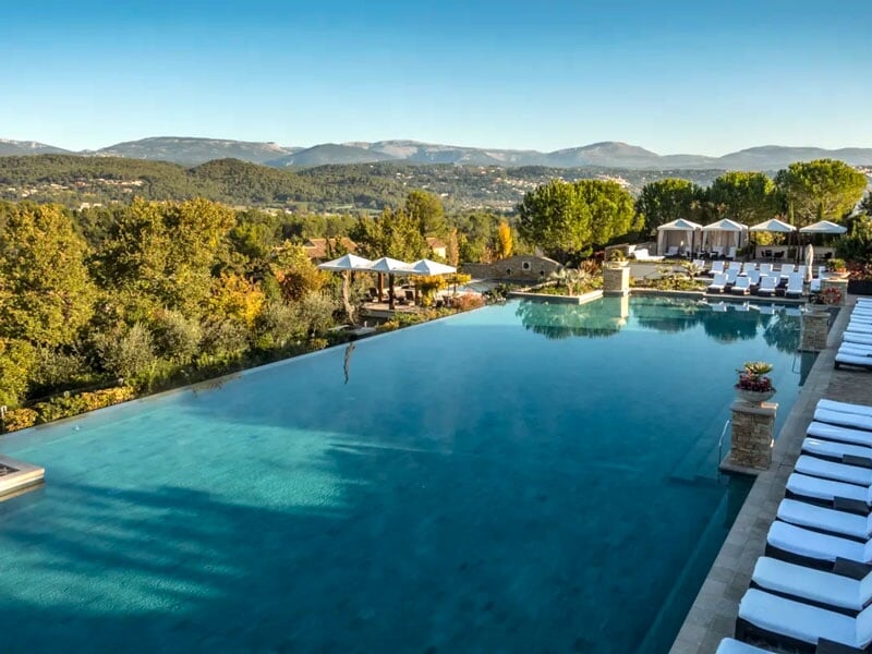 Piscine à débordement avec des chaises longues blanches, entourée d'arbres et offrant une vue sur les collines.
