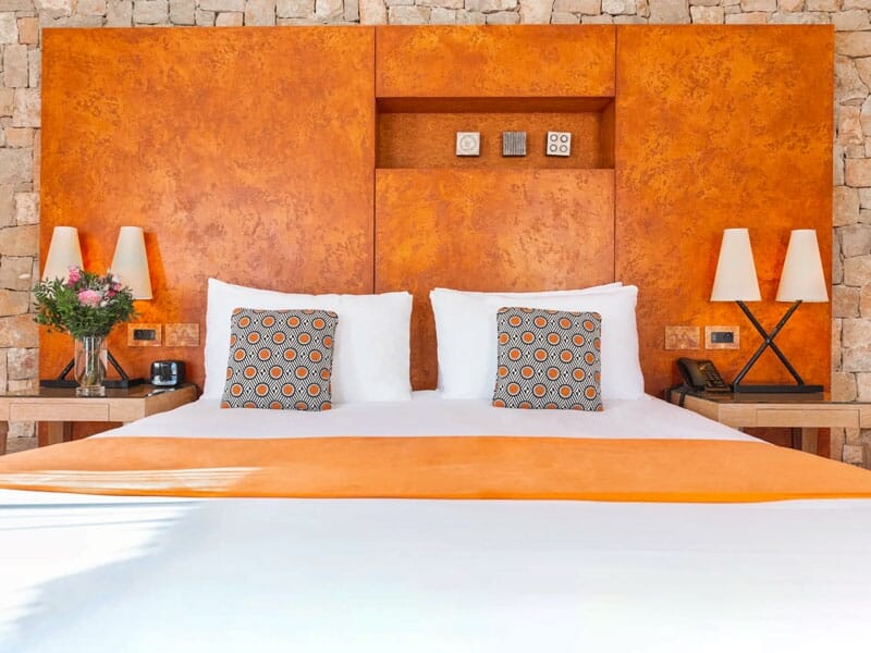 Chambre d'hôtel avec un grand lit, des coussins décoratifs orange, des lampes et un mur en pierre.