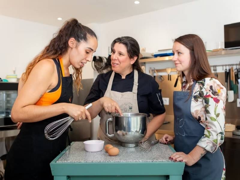 Trois femmes en tablier discutent et préparent une recette ensemble dans une cuisine moderne et bien équipée.