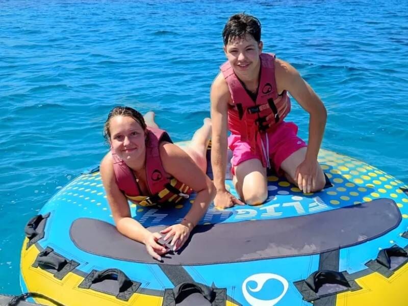 Deux adolescents posent sur une grande bouée bleue flottant sur une eau claire, tous deux portant des gilets de sauvetage.