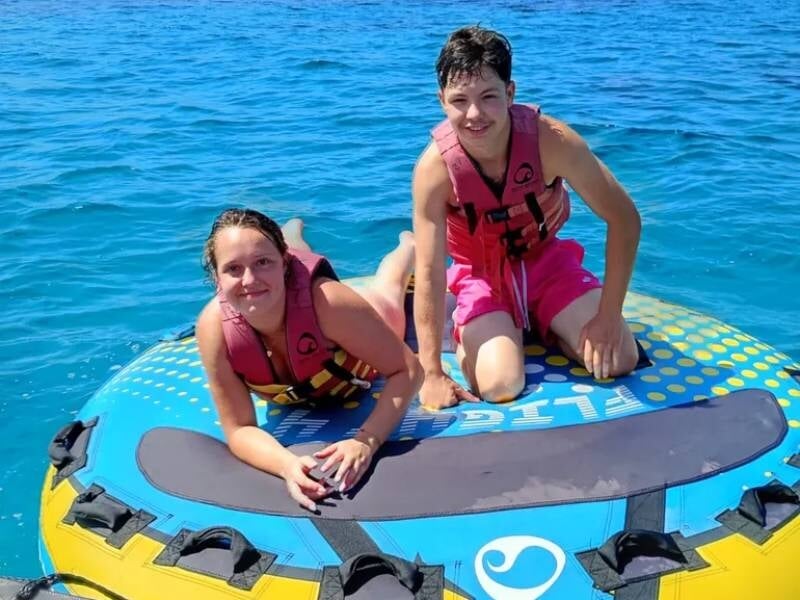 Deux adolescents posent sur une grande bouée bleue flottant sur une eau claire, tous deux portant des gilets de sauvetage.