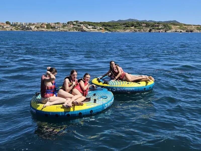 Quatre personnes en maillots de bain profitent d'une bouée tractée sur une mer calme avec un paysage côtier en arrière-plan.