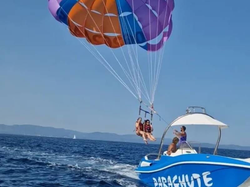 Deux personnes en parachute ascensionnel au-dessus de la mer, tirées par un bateau bleu avec un ciel clair en arrière-plan.