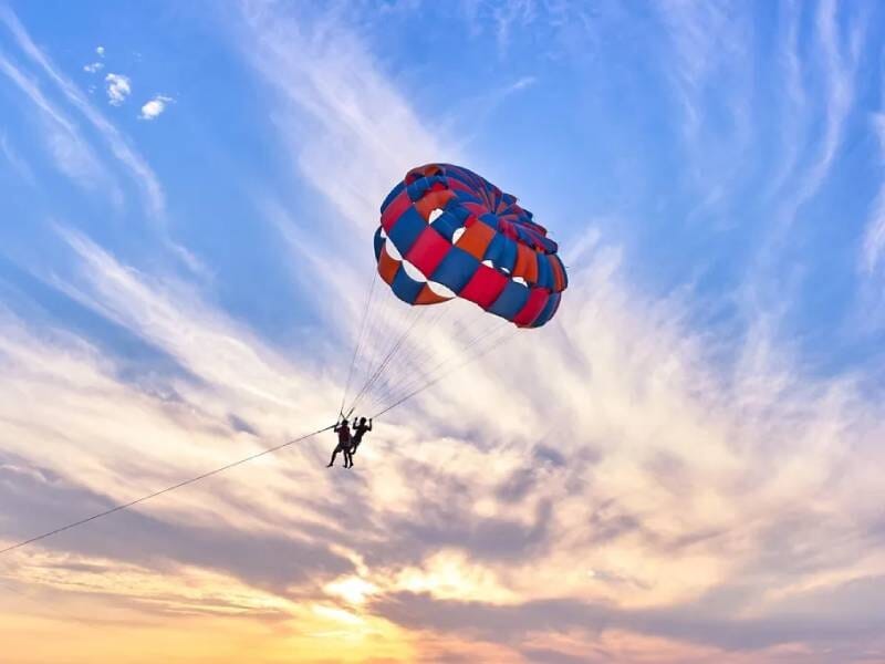 Deux personnes en parachute ascensionnel volant dans un ciel coloré au coucher du soleil, avec des nuages et un horizon visible.
