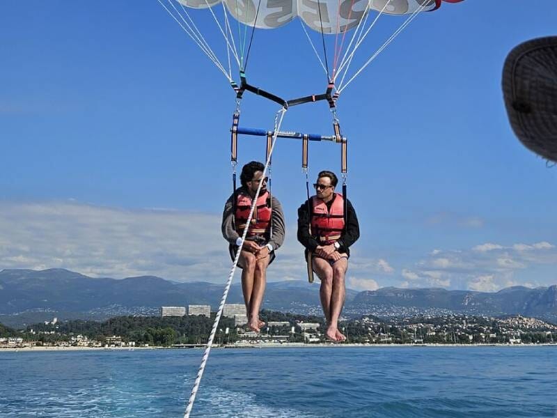 Deux hommes en gilets de sauvetage suspendus en parachute ascensionnel au-dessus de la mer, avec une côte et des collines en arrière-plan, à six-Fours-les-Plages.