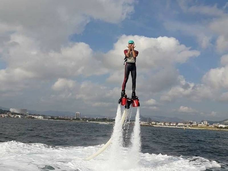 Homme en flyboard sur l'eau, entouré de jets d'eau, avec un ciel nuageux et une ville en arrière-plan.