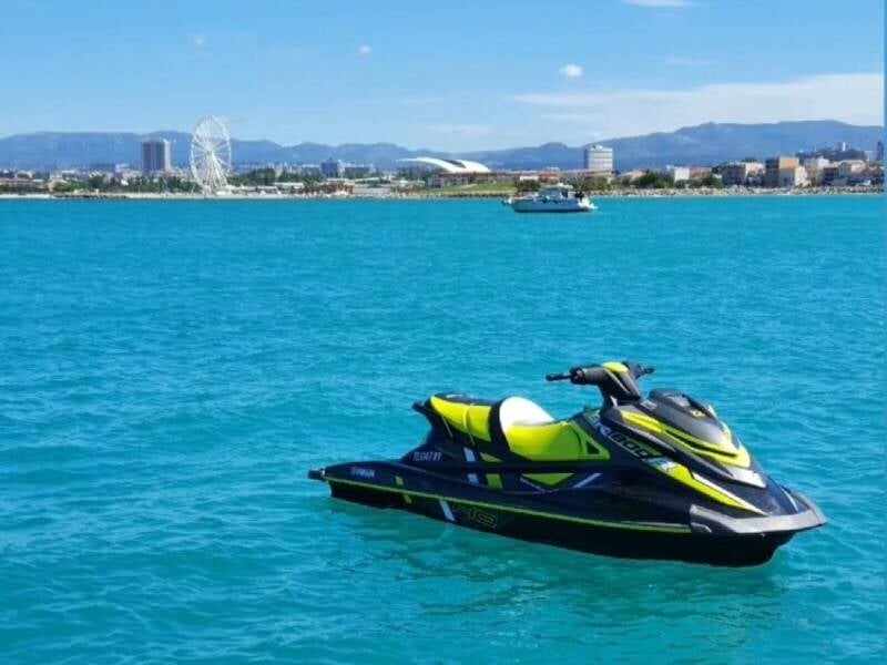Jet ski jaune et noir flottant sur une mer bleue avec une ville et une grande roue en arrière-plan sous un ciel clair.