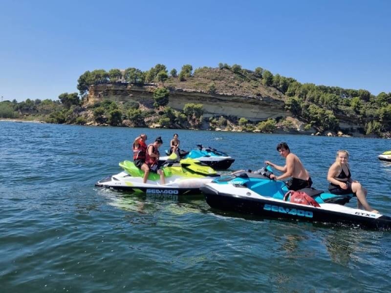 Quatre personnes sur des jet skis colorés sur une eau calme avec une falaise boisée en arrière-plan par temps ensoleillé.