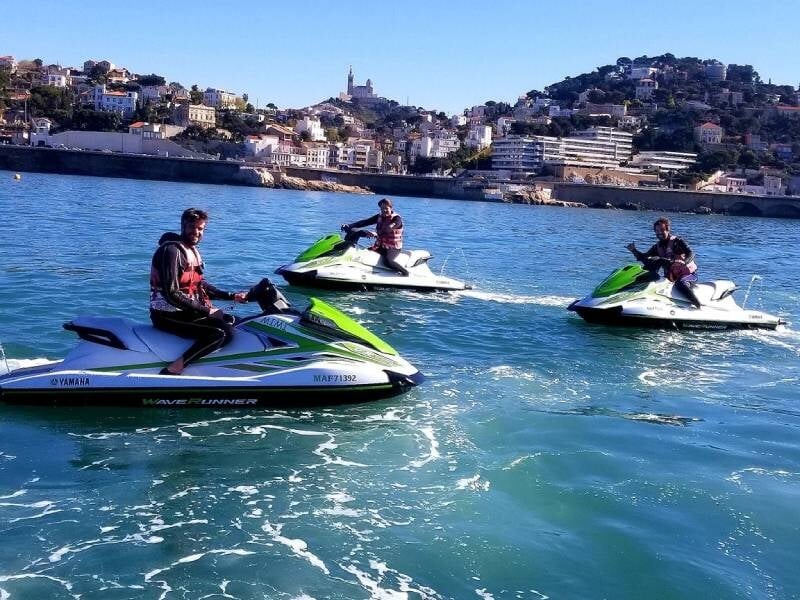 Trois personnes sur des jet skis verts et blancs naviguant sur une mer bleue avec une ville et une colline en arrière-plan, lors d'une randonnée près de Martigues.