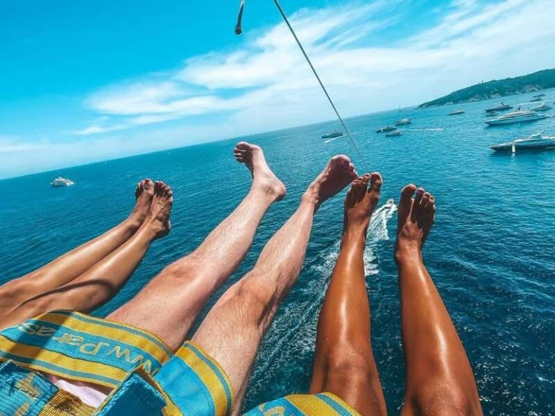 Vue des jambes de quatre personnes suspendues en parachute ascensionnel au-dessus de la mer et des bateaux, lors d'un vol à Ramatuelle.