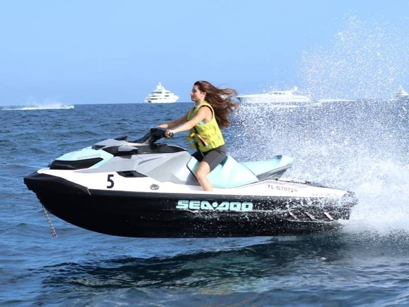 Jeune femme en gilet de sauvetage conduisant un jetski blanc et noir sur une mer agitée avec des bateaux en arrière-plan.