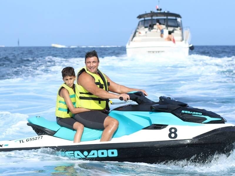 Homme souriant avec un enfant en gilet de sauvetage sur un jetski bleu et blanc, naviguant sur une mer calme avec un yacht derrière.