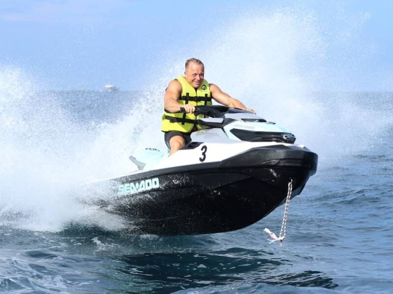 Homme en gilet de sauvetage conduisant un jetski noir et blanc, créant un grand éclaboussement d'eau sur une mer bleue lors d'une randonnée à Ramatuelle.