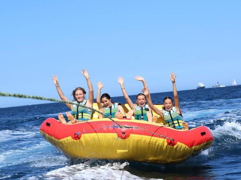 Quatre personnes souriantes sur une bouée tractée colorée rouge et jaune sur une mer bleue sous un ciel clair, à Ramatuelle.