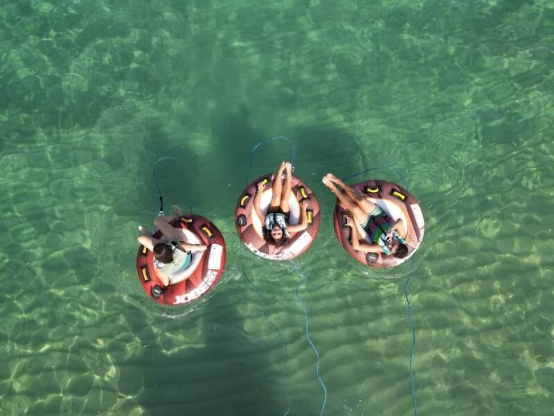 Trois personnes assises sur des bouées tractées rondes dans une eau claire et turquoise, vues de dessus.