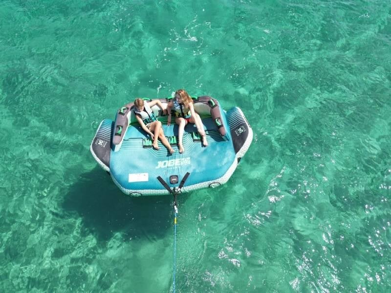 Deux personnes assises sur une grande bouée tractée bleue sur une mer turquoise, vues de dessus.