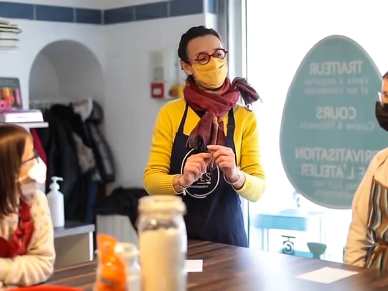 Femme en tablier et masque jaune expliquant une recette à un groupe dans une cuisine lumineuse et conviviale.