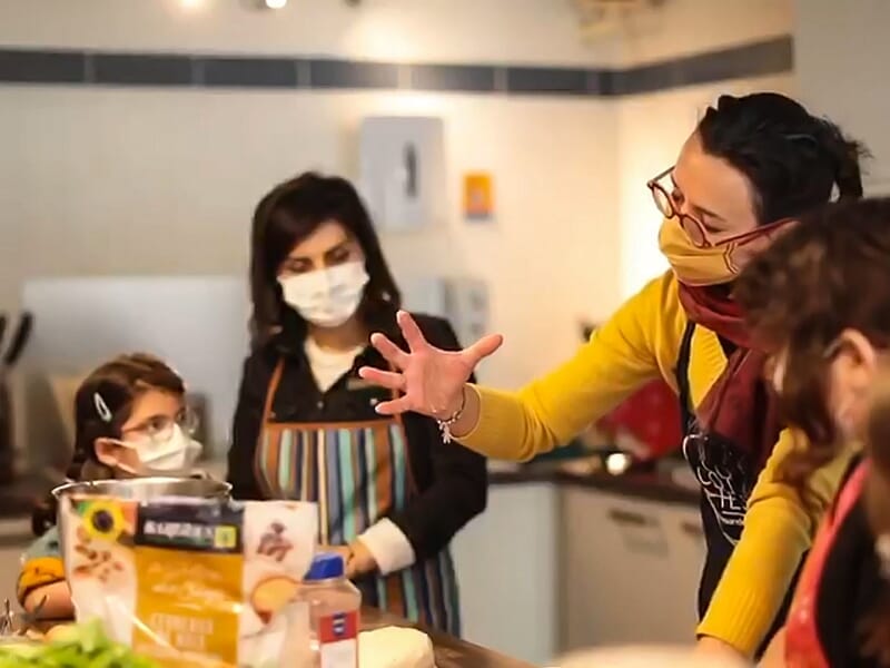 Instructrice en tablier et masque parlant à un groupe d'élèves portant des masques dans un cours de cuisine.