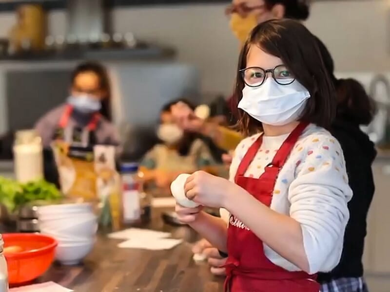 Jeune fille en tablier rouge et masque souriant en préparant un ingrédient dans une cuisine de groupe.