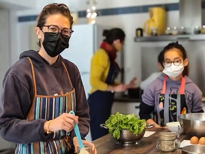 Personne en tablier rayé et masque noir tenant une spatule dans une cuisine avec d'autres participants.