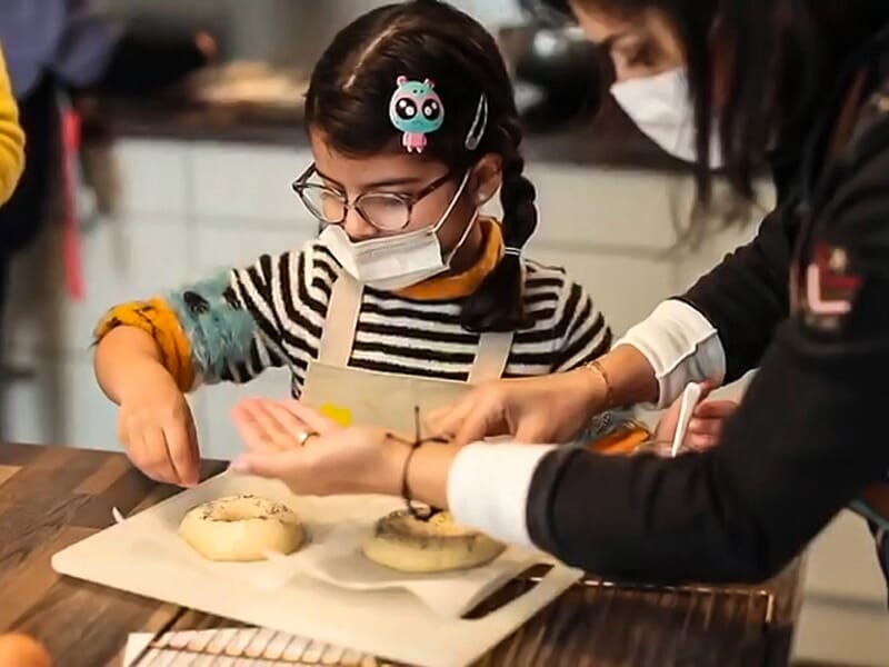 Enfant en tablier et masque décorant une pâte à pain sur une plaque de cuisson avec l'aide d'un adulte.