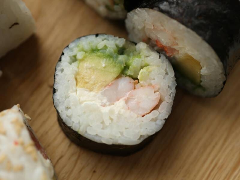 Gros plan sur un maki avec crevette, avocat et fromage frais, posé sur une surface en bois clair.