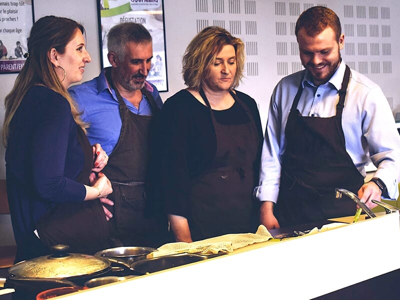 Groupe de quatre adultes en tabliers discutant et cuisinant autour d'une plaque de cuisson dans un atelier.