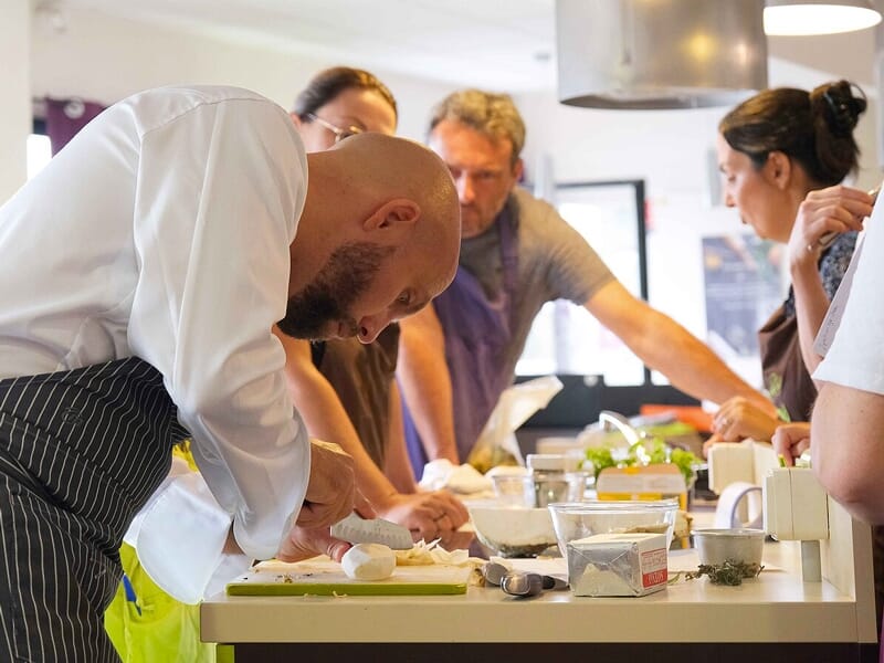 Chef cuisinier concentré découpe des ingrédients sur un plan de travail entouré de participants attentifs.