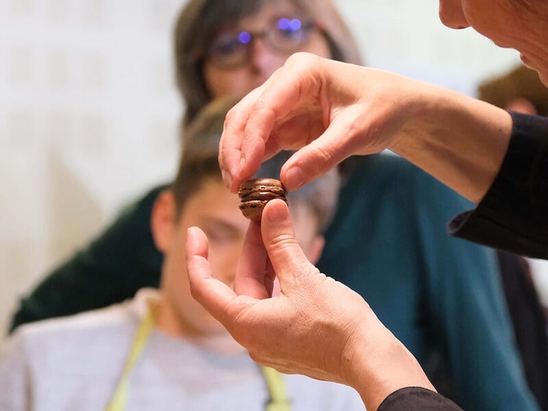 Main tenant un petit macaron chocolaté devant un groupe de personnes observant avec intérêt.