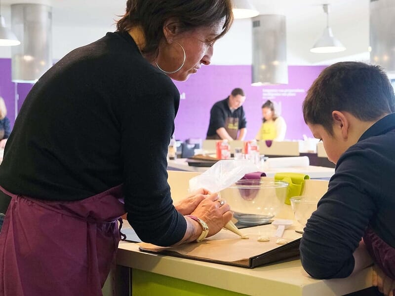 Femme en tablier montre à un garçon comment utiliser une poche à douille dans un atelier de pâtisserie.