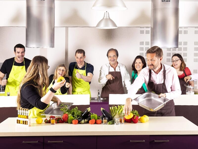 Groupe de huit personnes en tabliers souriants et cuisinant ensemble dans une cuisine équipée.