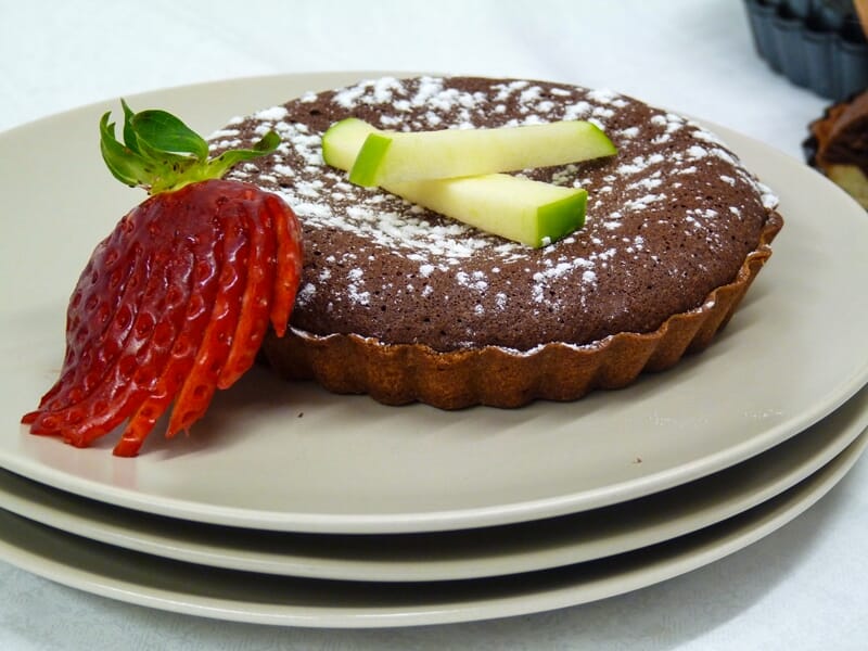 Tartelette au chocolat décorée de tranches de pomme verte et d'une fraise fraîche sur une assiette blanche.