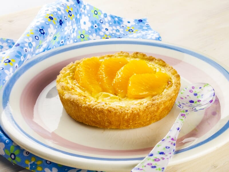 Tartelette aux quartiers d'orange sur une pâte dorée, servie avec une cuillère sur une assiette colorée.
