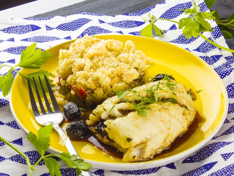 Filet de poisson blanc accompagné de couscous et olives noires sur assiette jaune avec herbes fraîches.