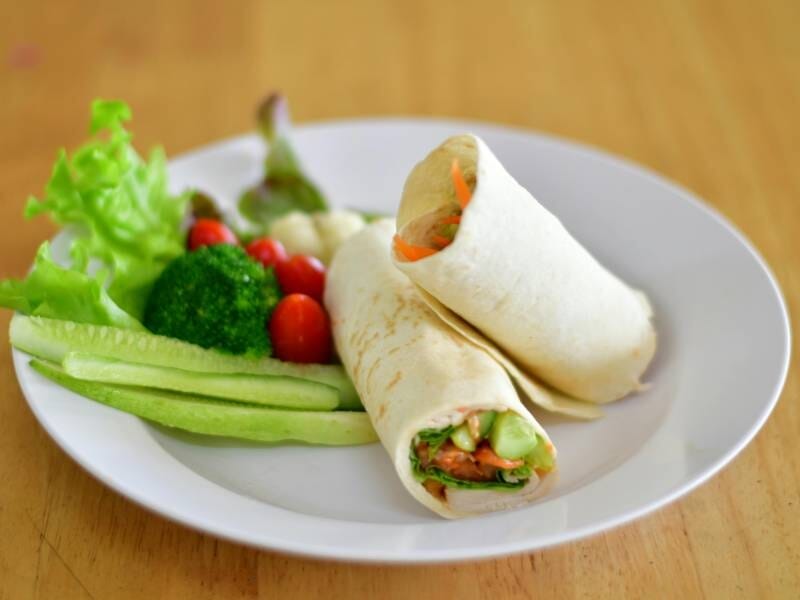 Deux wraps garnis de légumes frais accompagnés de brocoli, tomates cerises et bâtonnets de concombre sur une assiette blanche.