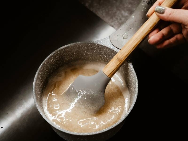 Une main remue une sauce épaisse dans une casserole grise avec une spatule en silicone à manche en bois.