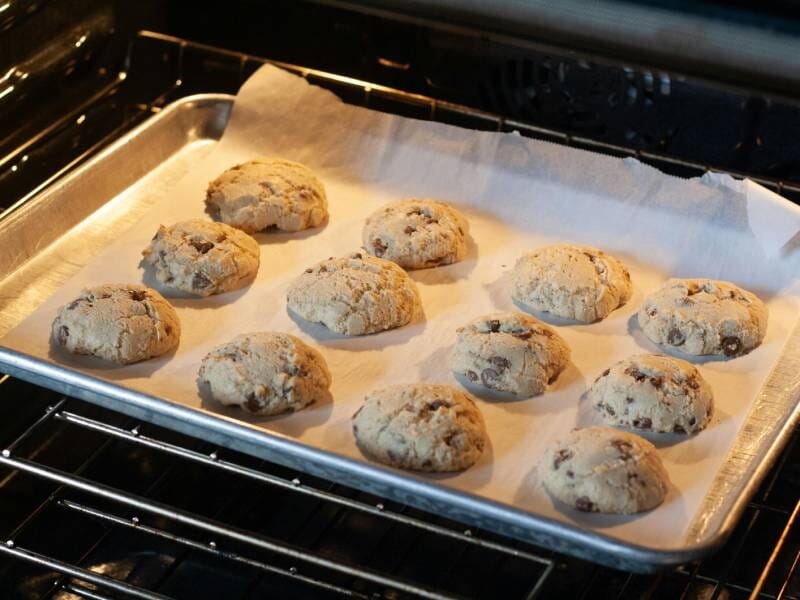 Des cookies aux pépites de chocolat cuisent sur une plaque recouverte de papier sulfurisé dans un four ouvert.