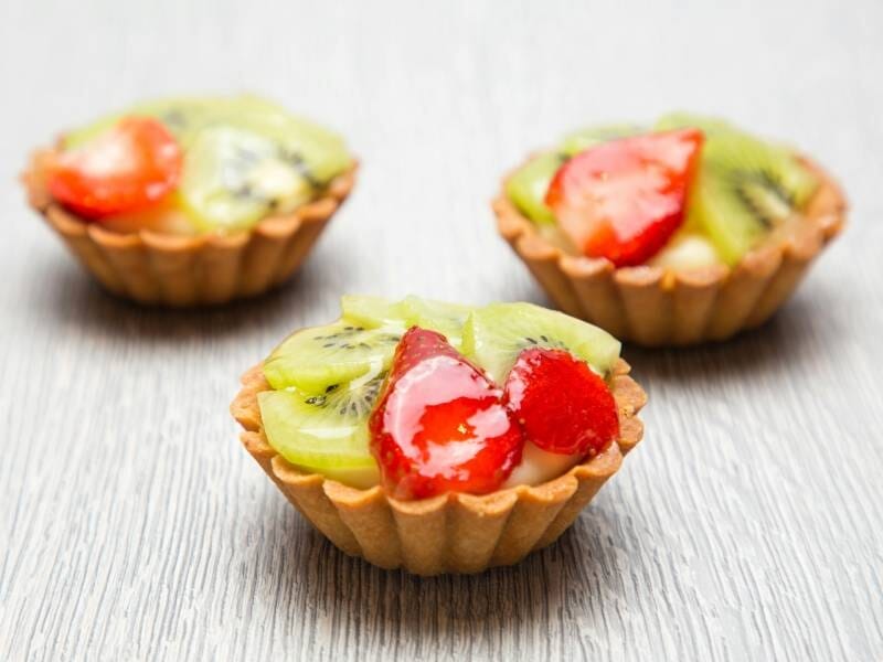 Trois petites tartelettes garnies de tranches de kiwi et de fraises sur une surface claire en bois, préparées lors d'un cours de pâtisserie à Dijon.