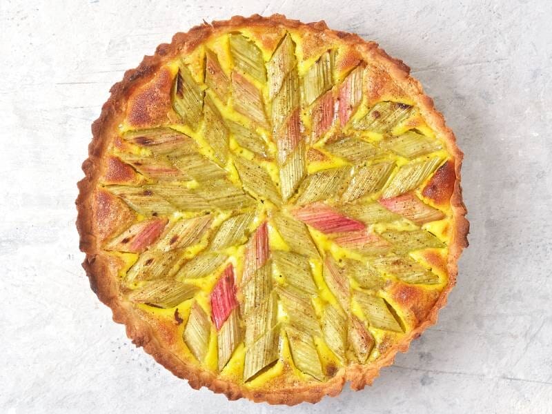 Tarte à la rhubarbe colorée disposée en motif géométrique sur une pâte dorée, posée sur une surface claire.