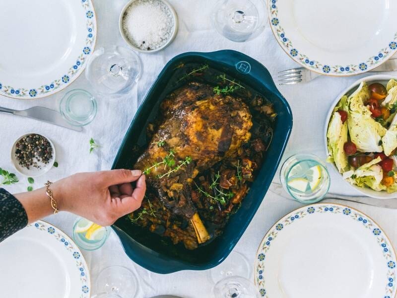 Plat de viande rôtie dans un plat bleu avec herbes fraîches, entouré d'assiettes et salade verte colorée.